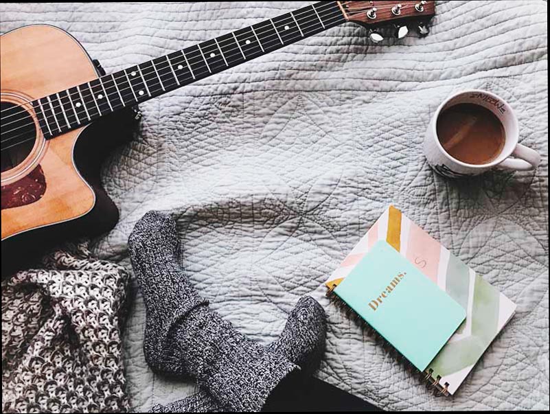 Foto de una persona sentada sobre una manta con una guitarra, un cuaderno y una taza de café.